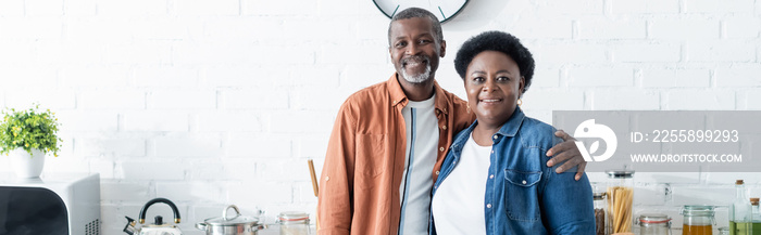 happy senior african american couple looking at camera in kitchen, banner.