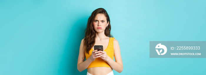 Confused and freak out young woman frowning, grimacing after reading strange message on smartphone, standing over blue background