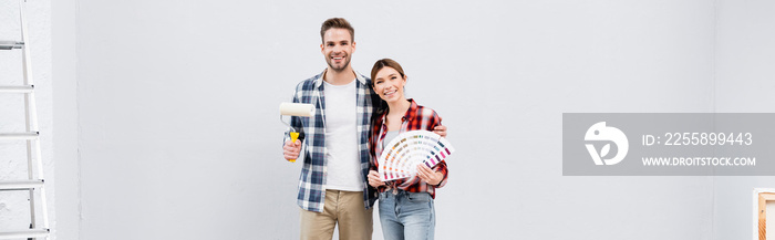 happy young man with paint roller hugging woman showing color palette at home, banner