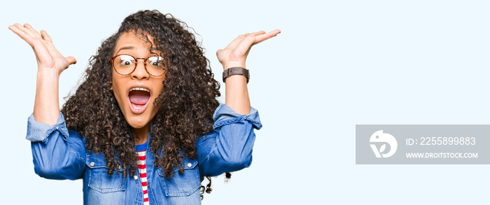 Young beautiful woman with curly hair wearing glasses celebrating crazy and amazed for success with arms raised and open eyes screaming excited. Winner concept