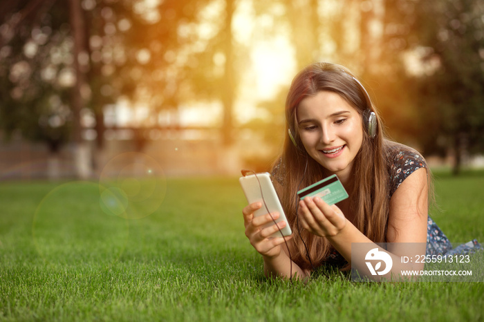 Portrait of a happy woman buying online with a smart phone outdoors lying down on green lawn headpho