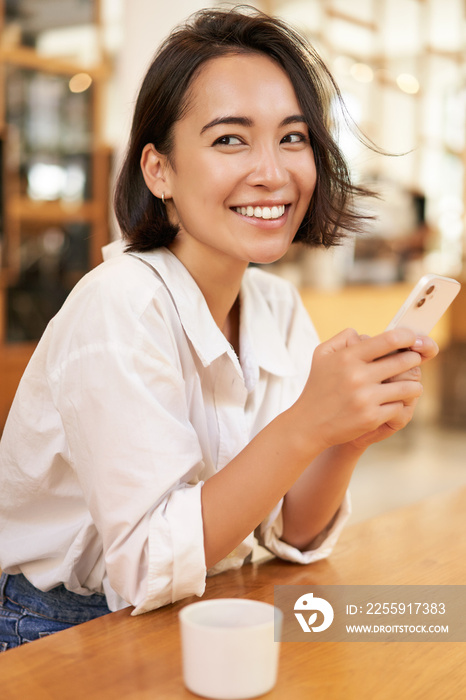 Vertical portrait of stylish asian woman sitting in cafe, drinking coffee and using smartphone