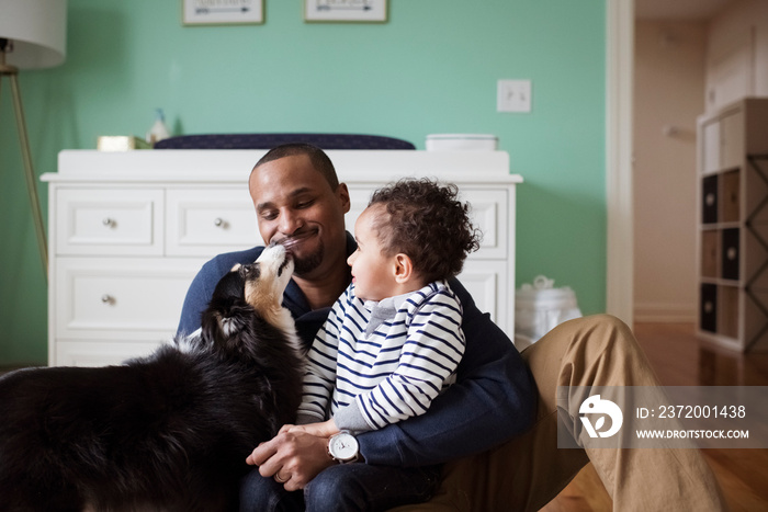 Dog licking father holding son while sitting against cabinet at home