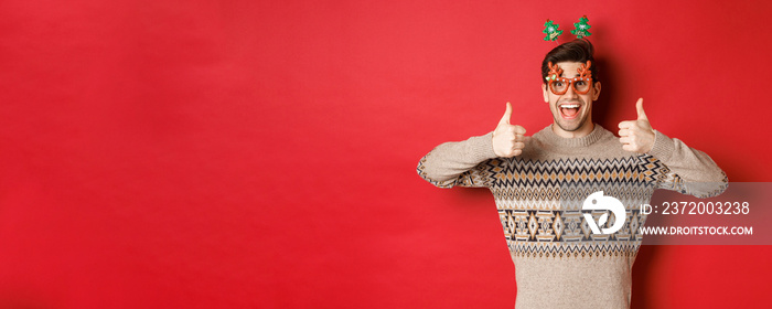 Portrait of satisfied and happy man in christmas sweater and party glasses, showing thumbs-up, wishi