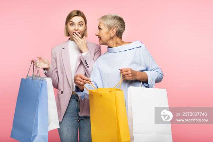 amazed young woman covering mouth near mature mother holding shopping bags isolated on pink