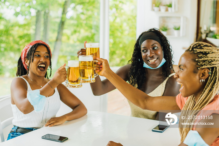 Three young African American girlfriends sitting at brewery (pub) with beer glasses taking off surgi