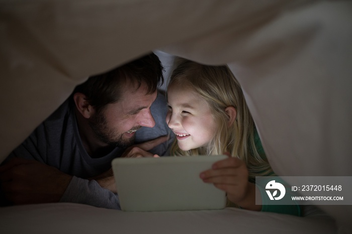 Father and daughter lying under blanket and using digital tablet