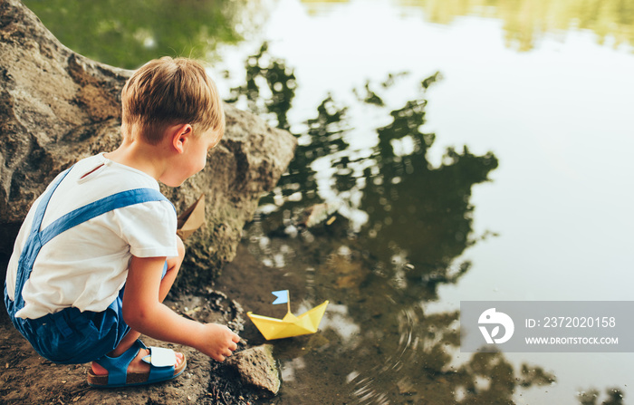 Rear view image of cute little boy launch paper ship on the lake in the park. Adorable kid boy playi