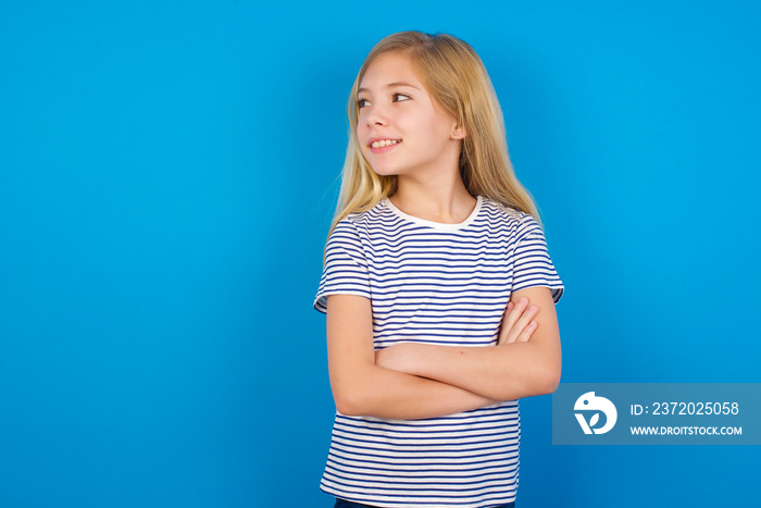 Dreamy rest relaxed Caucasian kid girl wearing striped shirt ​against blue wall crossing arms, looks