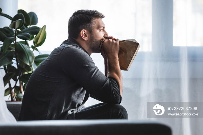 side view of handsome man with panic attack breathing in paper bag in apartment