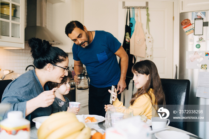 Girl showing smart phone to parents and brother during breakfast at home