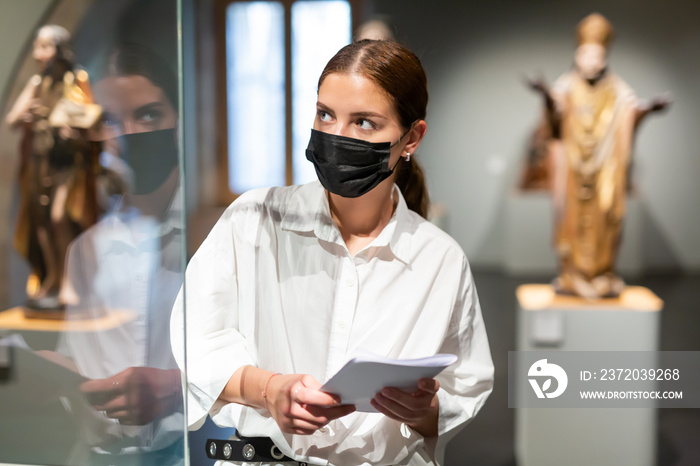 积极的年轻女性戴着防护面罩，带着纸质导游在画廊探索雕塑艺术作品