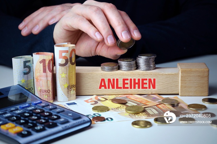 Anleihen. Mann stapelt Geld (Euro). Begriff Anleihen auf Baustein. Münzen, Scheine & Taschenrechner.