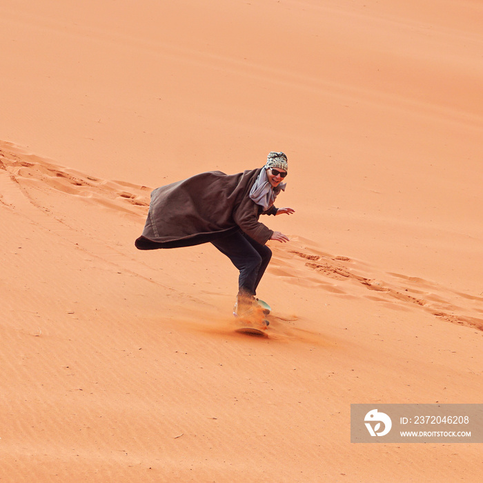 Young man smiling, sand dune surfing wearing bisht - traditional Bedouin coat. Sandsurfing is one of