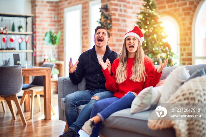 Young couple wearing santa claus hat sitting on the sofa around christmas tree at home crazy and mad
