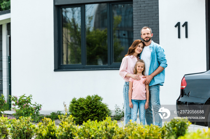 cheerful man standing with attractive wife and cute kid near new house