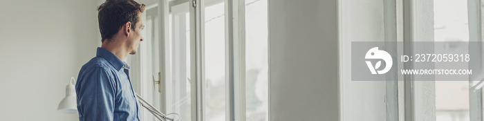 Wide view image of young businessman standing by bright office windows looking out.