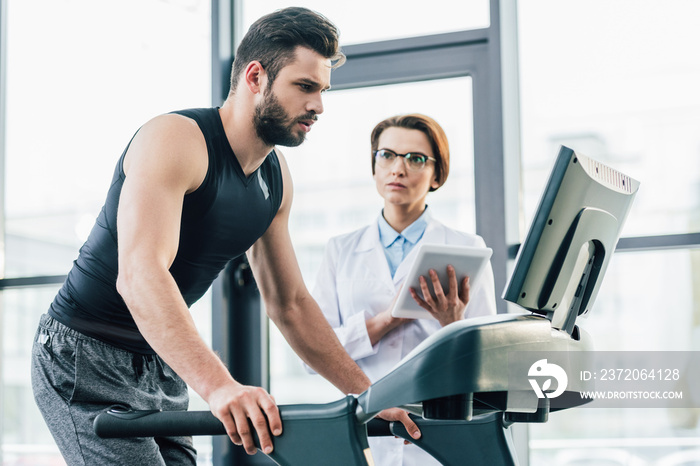 handsome sportsman running on treadmill near doctor during endurance test in gym