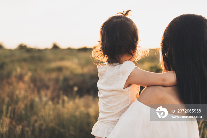 Cropped rear view of Caucasian brunette mother and child daughter embrace each other on meadow sunse