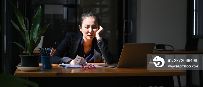 疲惫的年轻女商人拿着笔记本电脑坐在办公桌旁。