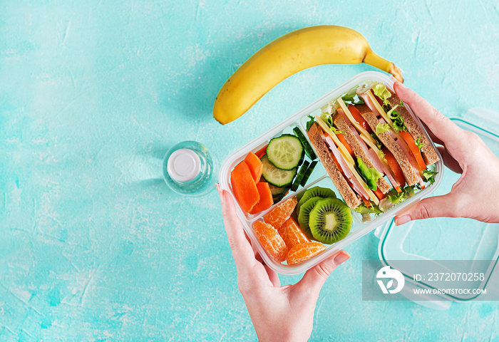 Lunchbox in hands. School lunch box with sandwich, vegetables, water, and fruits on table. Healthy e