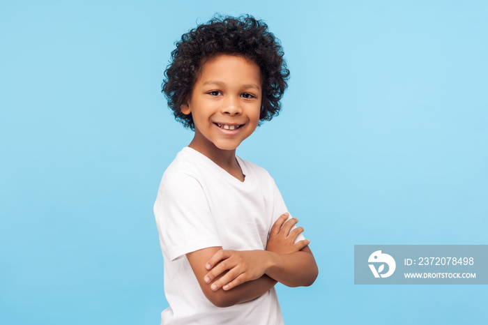 Portrait of happy and confident little boy in white T-shirt standing with crossed hands and smiling 