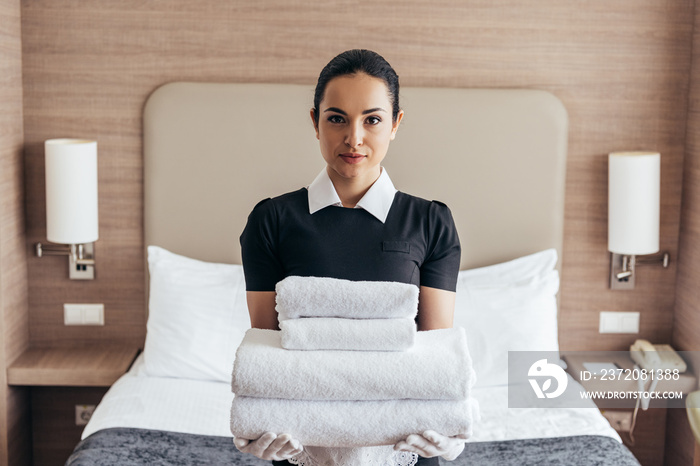 front view of maid holding pile of folded towels near bed and looking at camera in hotel room