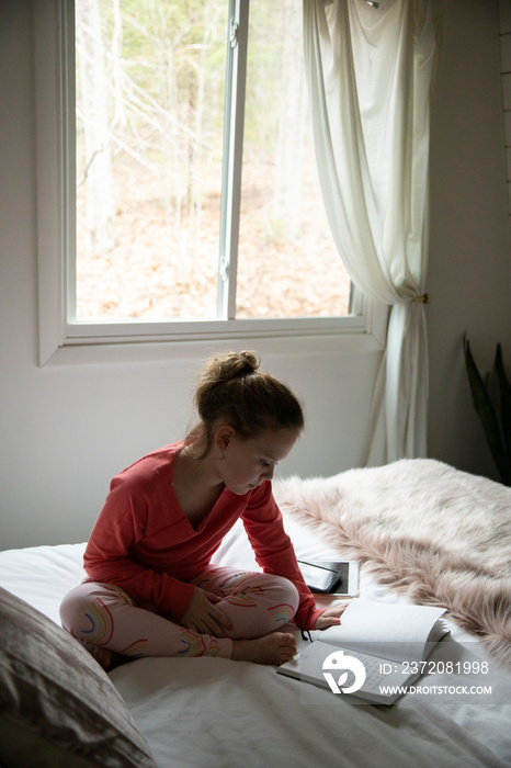 High angle view of girl reading book while sitting on bed against window at home