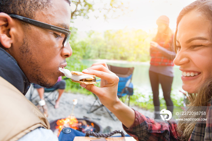 Young Adult Hispanic Girl Feeding African American Man A Smore at Campfire Outdoors