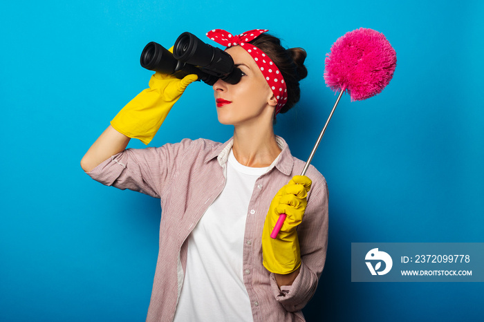 Smiling young woman in cleaning gloves holding dust brush and looking through binoculars on blue bac