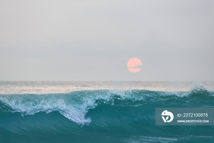 Blue ocean wave in front of beautiful sunset sky with pink sun disk over ocean skyline.