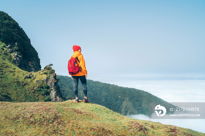 年轻的女背包客穿着橙色防水夹克在云层上方的山上徒步旅行