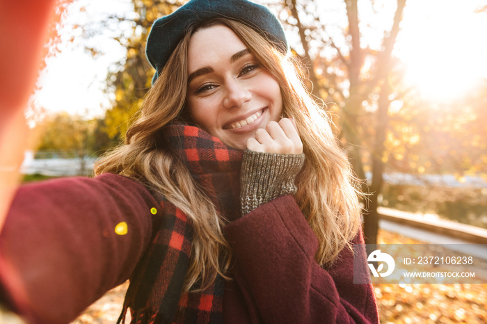 Beautiful happy young pretty woman walking outdoors in autumn spring park take a selfie by camera.