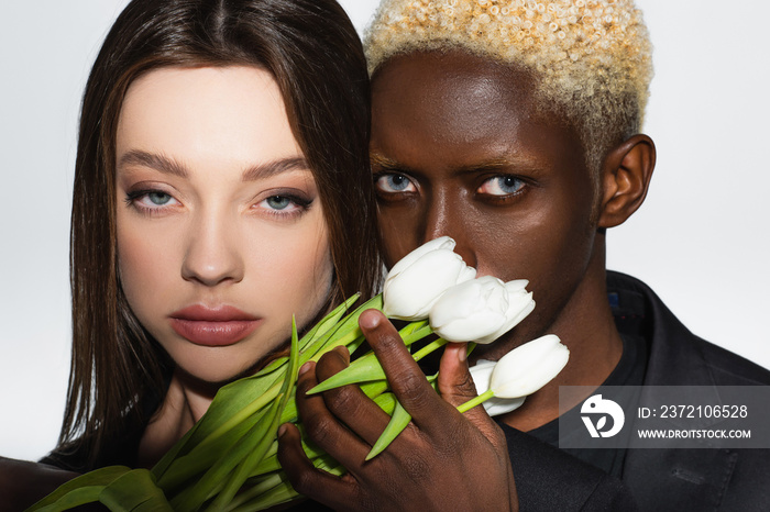 close up view of young interracial couple looking at camera near white tulips isolated on grey.