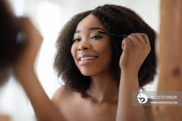 Gorgeous black woman applying mascara on her lashes in front of mirror at home