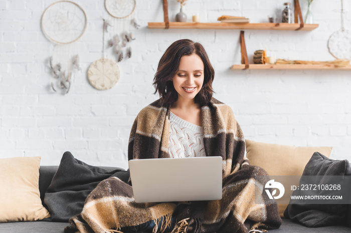 beautiful cheerful girl in blanket using laptop in living room with dream catchers