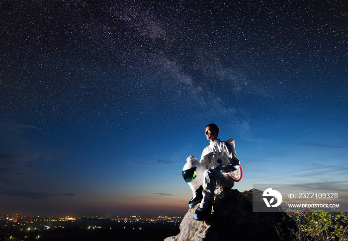 宇航员坐在岩石山顶，看着美丽的夜空的侧视图。太空人wea