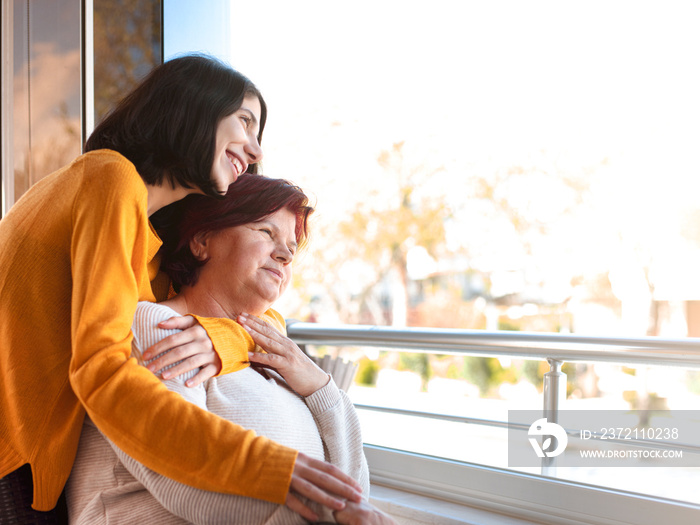 Happy senior mother and adult daughter looking at the view through the window.