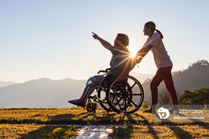 Disabled handicapped young man in wheelchair walking with his care helper in sunset.Silhouette