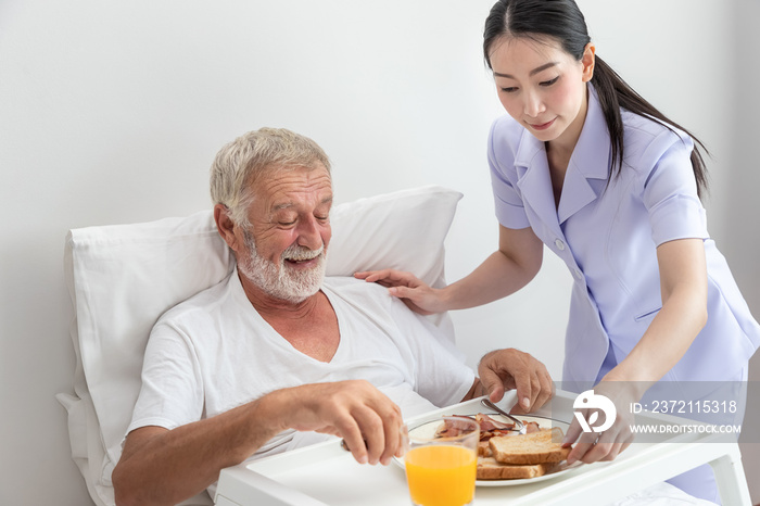 Happy nurse assist and help elderly senior man to eat breakfast on bed in bedroom at nursing home