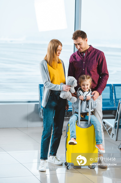 adorable child sitting on suitcase while mother holding teddy bear and standing near husband in wait