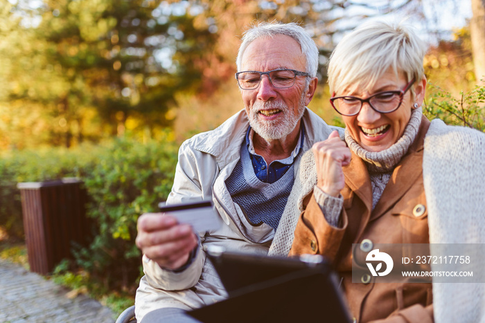 Senior couple sitting in public park using smart device to shop online. Cheerful seniors using moder