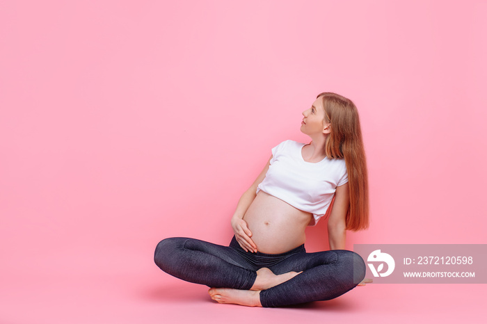 young pregnant girl sitting on a pink background, looking to the side of empty space