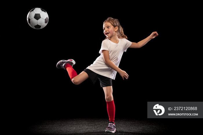 黑色背景下，顽皮的小女孩踢着球，双脚分开的全长肖像