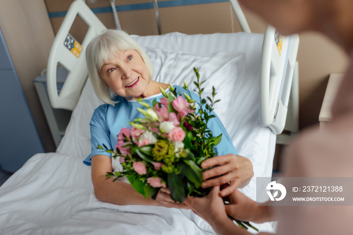 daughter presenting flowers to happy senior woman lying in bed in hospital