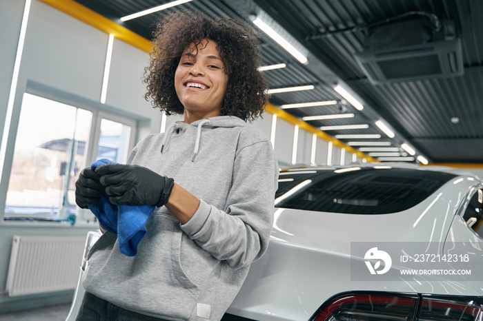 Cheerful detailer with microfiber towel leaning back against trunk lid