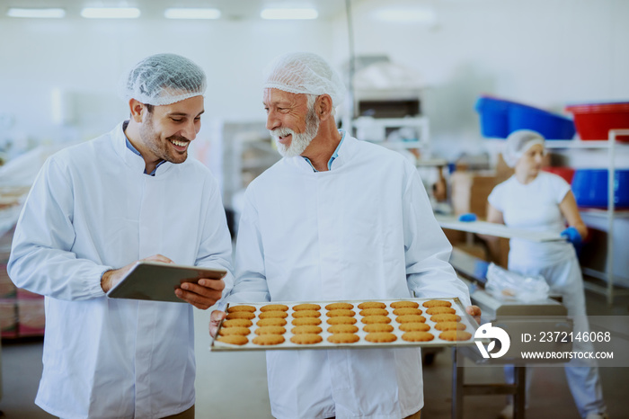 Senior adult employee in sterile white uniform standing with tray with cookies in food plant. Next t