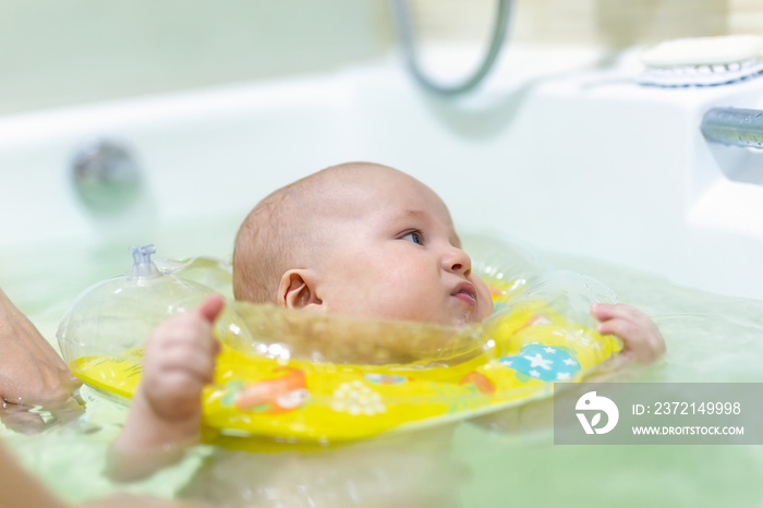 Cute little infant boy swimming with inflatable neck swim ring in bath. Baby having fun during water