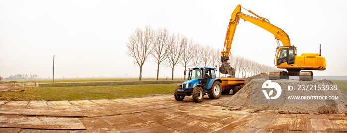 Crane filling a tipper trailer with sand, pulled by a tractor, in a misty polder landscape in winter