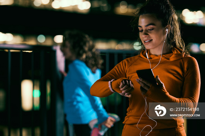 A young smiling night runner standing outdoors at night, using a smart phone and looking at a smart 